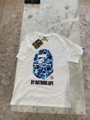  BAPE t-shirt  - 100% BOMULL. Säljer en vit t-shirt från BAPE med det ikoniska apansikte-trycket i blått kamomönster på bröstet och texten 'BY BATHING APE' under. Klassisk passform och rund hals. Perfekt för dig som gillar streetwear och vill sticka ut med en unik design.