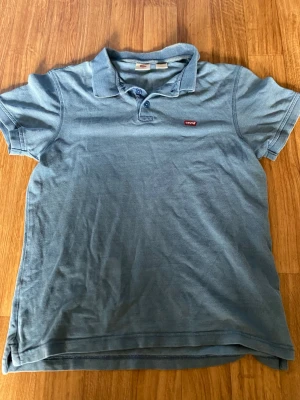 Blå pikétröja från Levi's - Säljer en klassisk blå pikétröja från Levi's med korta ärmar och krage. Tröjan har knappar vid halsen och den ikoniska röda Levi's-loggan på bröstet. Perfekt till jeans eller shorts för en avslappnad look.