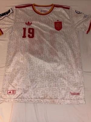 Vit Spanien matchtröja Adidas - Säljer en vit Spanien matchtröja från Adidas med röda detaljer, nummer 19 och gulröd krage. Tröjan har ett diskret mönster, broderad landslagssköld och Adidas-logga på bröstet. Perfekt för fotbollsfans som vill sticka ut på läktaren eller planen.