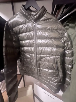 Olivgrön dunjacka från Moncler - Snygg olivgrön dunjacka från Moncler med quiltad design och hög krage. Jackan har dragkedja framtill, två fickor med dragkedja och klassisk Moncler-logga på ärmen. Perfekt för kalla vinterdagar och riktigt lätt att bära.