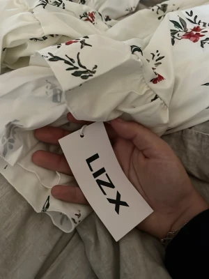 Vit blommig klänning från LIZZ - Söt vit klänning från LIZZ med små röda och gröna blommor, volangdetaljer framtill. Klänningen har lång ärm och smock i midjan för en bekväm passform. Perfekt för vår och sommar!