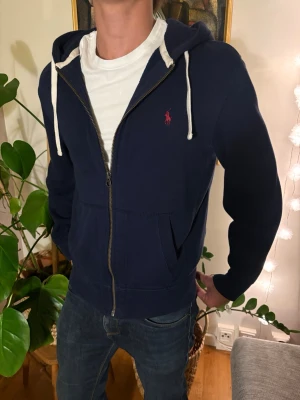 Ralph lauren zip - Mörkblå hoodie med vit dragsko och dragkedja framtill. Klassisk passform och fickor på sidorna. Liten röd logga broderad på bröstet. Perfekt till jeans och enkel att matcha. Mjuk och skön känsla tack vare bomullsmaterial. Storlek m