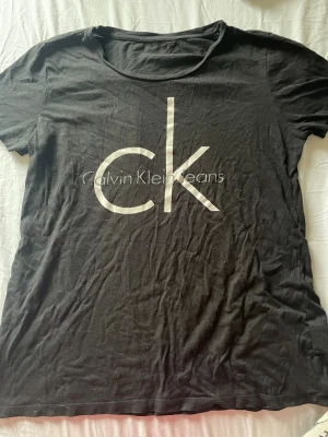 Svart Calvin Klein t-shirt - Snygg svart t-shirt från Calvin Klein Jeans med stort vitt ck-tryck på bröstet. Klassisk rund hals och korta ärmar. Tillverkad i mjuk bomull som känns skön mot huden. Perfekt för en clean och stilren look.