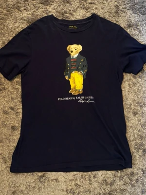 Svart Polo Bear t-shirt från Ralph Lauren - Står Xl men passar som S. Svart t-shirt från Ralph Lauren med ikoniska Polo Bear-trycket på bröstet. Björnen har på sig en randig tröja, kavaj och gula byxor. T-shirten har rund hals och korta ärmar, perfekt för en avslappnad och stilren look.