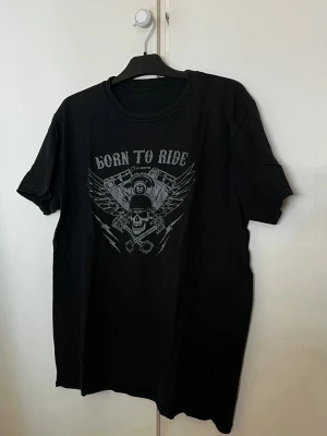 Svart MC  t-shirt Born To Ride - Cool svart t-shirt med trycket 'Born To Ride' och motiv av dödskalle, motor och vingar på bröstet. Klassisk rund hals och korta ärmar. Perfekt för dig som gillar bikerstil och vill sticka ut.