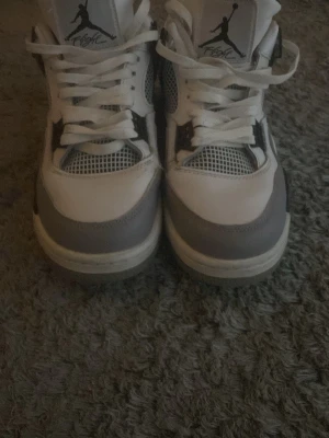 Vita och gråa Air Jordan 4 sneakers - Snygga Air Jordan 4 sneakers i vitt och grått med svarta detaljer. Skorna har klassisk mesh på sidorna, högt skaft och ikoniska Jumpman-loggan på plösen. Perfekta för dig som gillar streetwear och vill ha en tidlös look.