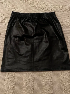 Svart kortkjol i skinnimitation - Säljer en svart kortkjol i skinnimitation med elastisk midja. Kjolen har en enkel och stilren design som ger en cool vibe till din outfit. Perfekt att matcha med en stickad tröja eller crop top för en trendig look. Lappen borta vet inte vart den är ifrån