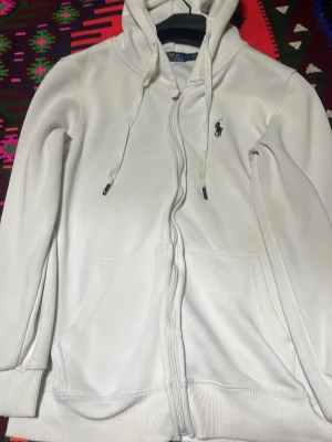 Vit hoodie med dragkedja från Polo Ralph Lauren - Vit hoodie från Polo Ralph Lauren med dragkedja framtill, justerbar huva med snören och två stora fickor. Klassisk mörkblå logga broderad på bröstet. Tillverkad i mjuk bomull för en skön och avslappnad look.