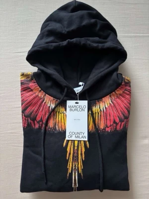 Marcelo Burlon Hoodie - Svart hoodie från Marcelo Burlon med ikoniskt färgglatt vingmotiv i rött, orange och gult över axlar och bröst. Klassisk huva med dragsko och stor magficka framtill. Perfekt för dig som vill sticka ut med en unik och trendig look. Pris kan diskuteras privat