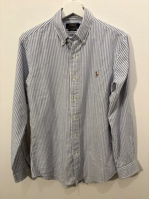Randig skjorta från Polo Ralph Lauren - Klassisk skjorta från Polo Ralph Lauren med blå och vita ränder. Skjortan har lång ärm, knappar framtill och en liten broderad logga på bröstet. Tillverkad i bomull och har en normal passform. Perfekt för en clean och stilren look. 