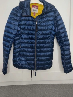 Blå pufferjacka med huva Parajumpers - Snygg blå pufferjacka från Parajumpers med huva och dragkedja framtill. Jackan har quiltad design, två sidofickor med dragkedja och gul insida vid kragen. Perfekt för kalla vinterdagar och riktigt lätt att bära.