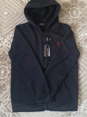 Svart hoodie från Polo Ralph Lauren - Snygg svart hoodie från Polo Ralph Lauren med dragkedja och huva. Klassisk röd logga broderad på bröstet. Tillverkad i mjukt bomullsmaterial och har två fickor framtill. Perfekt för en avslappnad och stilren look.