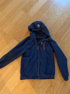 Blå hoodie från Sail Racing - Mörkblå hoodie från Sail Racing med dragkedja framtill, huva och en bröstficka med dragkedja. Snygg logga på bröstet och huvan. Perfekt för chill dagar eller när du vill ha en sportig look.