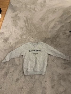 Grå sweatshirt från Björn Borg - Snygg grå sweatshirt från Björn Borg med svart text på bröstet. Tröjan har rund halsringning, ribbade muddar vid ärmslut och nederkant samt en klassisk passform. Perfekt för en avslappnad och sportig stil.