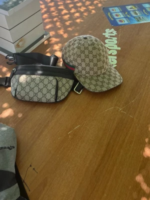 Beige keps från Gucci med GG-mönster BÅDA SÄLJS! - Säljer en beige keps från Gucci med det klassiska GG-monogrammönstret över hela kepsen. Kepsen har en böjd skärm och en grön-röd rand på sidan. Perfekt accessoar för dig som gillar exklusiv och trendig streetwear. Med kepsen går de med att köpa med väskan oxå!
