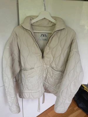 Beige quiltad pufferjacka från Zara - Säljer min trendiga beige quiltad pufferjacka från Zara med dragkedja framtill, hög krage och två stora fickor. Jackan har vågigt mönster i sömmarna och dragsko i nederkant för justerbar passform. Perfekt nu till hösten!