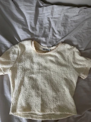 Vit spetsblus med kort ärm - Söt beige topp i spets med korta ärmar och rund hals. Blusen har ett fint spetsmönster över hela plagget och är lätt transparent. Perfekt att styla med jeans eller kjol för en trendig look.