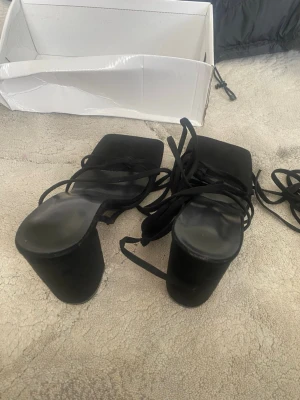Svarta platåsandaler med snörning - Säljer ett par svarta sandaler med hög platåklack och fyrkantig tå. Sandalerna har tunna remmar som snöras runt foten och benet för en trendig look. Materialet ser ut att vara mockaimitation och de har en chunky klack som ger extra höjd. Stl 36