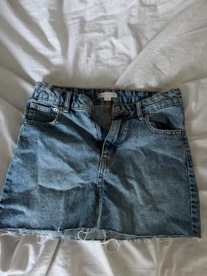 Kort jeanskjol med rå kant - Säljer en klassisk blå jeanskjol i kort modell med rå, fransig kant nedtill. Kjolen har fem fickor, bälteshällor och dragkedja framtill. Perfekt till sommaren och enkel att styla med olika toppar.