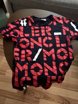 Svart moncler t-shirt med röd text - Snygg svart moncler t-shirt med stora röda bokstäver och vita detaljer över hela tröjan. T-shirten har rund hals och korta ärmar. Perfekt för dig som gillar grafiska tryck och vill sticka ut lite extra.