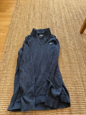 Mörkblå halvzip tröja Hollister - Mörkblå stickad tröja från Hollister med halvzip och ribbad krage. Tröjan har långa ärmar, ribbade muddar och en liten vit logga på bröstet. Perfekt för lager på lager och en avslappnad stil.