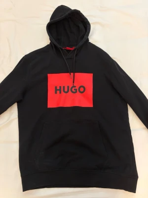Svart hoodie med röd HUGO-logga - Svart hoodie från HUGO med stor röd logga på bröstet. Klassisk huva med dragsko och stor magficka framtill. Tillverkad i mjukt material som passar perfekt för en avslappnad stil. Ribbade muddar vid ärmslut och nederkant.