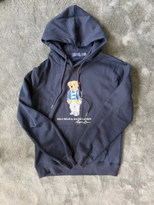 Mörkblå hoodie Polo Bear Ralph Lauren - Mörkblå hoodie med huva och dragsko från Ralph Lauren. På bröstet finns ett tryck av den ikoniska Polo Bear i randig tröja och jacka. Mjuk bomullskänsla och ribbade muddar. Perfekt för en avslappnad och trendig stil.