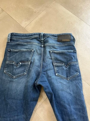 Blå slim fit jeans från Diesel - Säljer ett par blå slim fit jeans från Diesel med snygg tvätt och slitningar. Jeansen har klassiska fem fickor, raka ben och detaljerade bakfickor med V-formad söm. Tillverkade i denim med skön stretch för extra komfort.