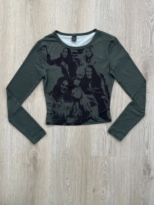 Mörkgrön croppad långärmad topp - Trendig mörkgrön croppad topp med lång ärm och tryck av flera personer på framsidan. Toppen har en slim passform och är tillverkad i ett stretchigt syntetmaterial. Perfekt för dig som vill sticka ut med ett coolt motiv.