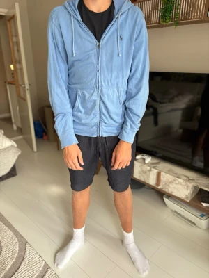 Ljusblå zip hoodie med huva - Ljusblå hoodie med dragkedja framtill och två stora fickor. Tröjan har huva med dragsko och ribbade muddar vid ärmslut och nederkant. Sitter som L/XL.
