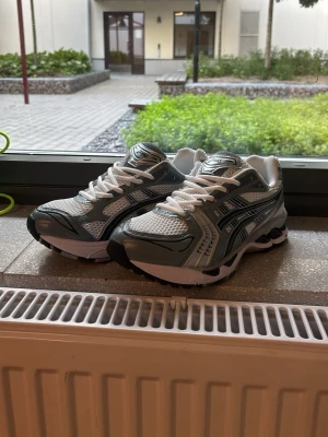 Asics  sneakers - Allt OG! Säljer ett par fräscha Asics sneakers i silver och vitt med svarta detaljer. Skorna har en sportig design med mesh-ovandel för bra ventilation och vita snören. Sulan är tjock och ger bra stöd, perfekt för dig som gillar streetwear och sportig stil.