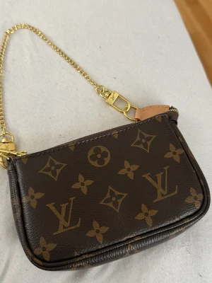 Louis Vuitton mini pochete  - Säljer en brun plånbok från Louis Vuitton med klassiskt monogrammönster och guldfärgad kedja. Plånboken har dragkedja och detaljer i läder samt metall. Perfekt för kort och småsaker. Har inga kvitton eller låda kvar 