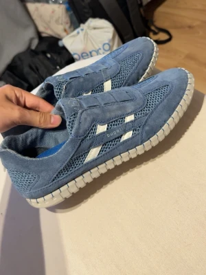 Blå sneakers från Bugatti - Snygga blå sneakers från Bugatti med vita detaljer och grov sula. Ovandelen är i mocka och mesh för en sportig look och bra ventilation. Skorna har slip-on design utan snörning och vita ränder på sidan. Perfekta för en avslappnad streetstyle. De har använts 3 gånger totalt.