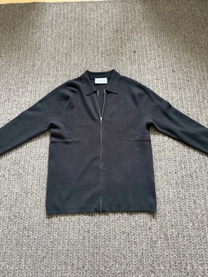 Svart zip-cardigan från Ciszere - Stilren svart cardiganjacka från Ciszere med dragkedja framtill och klassisk krage. Tillverkad i mjukt stickat material som ger en clean look. Perfekt att slänga över en t-shirt för en enkel men snygg outfit.