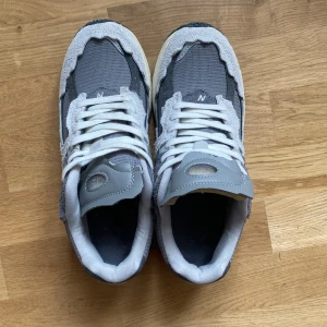 Grå New Balance 2002R sneakers - Snygga New Balance 2002R sneakers i olika grå nyanser med vit sula och vita skosnören. Ovandelen är i mesh och mocka med tydliga lager och den klassiska N-loggan på sidan. Skorna har en chunky sula och sportig vibe, perfekta för dig som gillar streetwear.