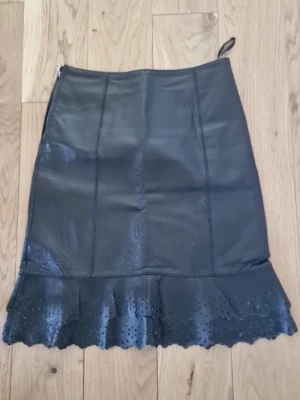 Svart läderkjol från vero moda - Snygg svart läderkjol från Vero Moda med dekorativ volangkant nertill och hålbroderade detaljer. Kjolen har en rak passform och dragkedja i sidan. Perfekt för dig som vill ha en edgy look. Jätte fin kjol som är knappt använd i väldigt bra skick, passar storlek s/xs.💗