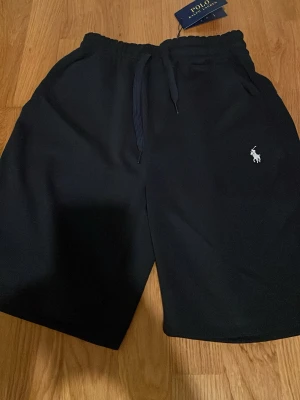 Mörk blåa shorts från Polo Ralph Lauren - Mörk blåa shorts från Polo Ralph Lauren och klassisk logga broderad på vänster ben. Tillverkade i mjukt material som passar perfekt för avslappnade dagar. Enkel och stilren design med elastisk midja.