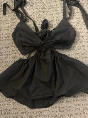 Svart topp med knyt och omlott - Säljer en svart omlott topp med knyt i ryggen och runt nacken. Toppen har en unik design med öppen rygg och voluminös nederdel. Perfekt för dig som vill sticka ut med en trendig och cool look.