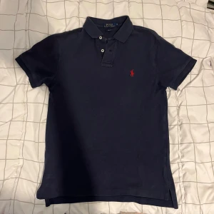 Marinblå pikétröja från Polo Ralph Lauren - Klassisk marinblå pikétröja från Polo Ralph Lauren med korta ärmar, krage och knappar framtill. Tröjan har den ikoniska röda logotypen broderad på bröstet och är tillverkad i mjuk bomull för en bekväm känsla.