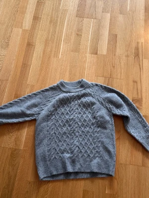 Grå stickad tröja med mönster - Mysig grå stickad tröja med ett snyggt diamantmönster framtill. Tröjan har rund halsringning, långa ärmar och ribbade muddar vid ärmslut och nederkant. Perfekt för kyliga dagar och enkel att matcha med jeans eller kjol.