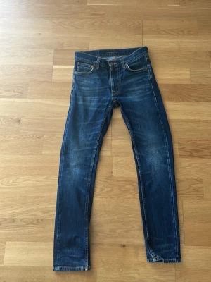 Blå lean Dean nudie jeans med slitningar - Säljer ett par blå nudie jeans med snygga slitningar och klassisk femficksdesign. Jeansen har normalhög midja och smala ben. Materialet är denim i bomull med lite stretch för skön passform. Perfekta till en avslappnad stil. Har knappt använts nåt