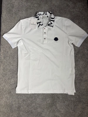 Monclertshirt - Vit pikétröja med korta ärmar och klassisk krage med stor logotyptext. Tröjan har knappslå framtill och en diskret svart på bröstet. Tillverkad i mjuk bomull för en clean och stilren look.
