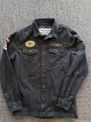 Svart långärmad skjorta Superdry - Svart långärmad skjorta från Superdry Rookie Edition med militärinspirerade patches på bröstet och ärmen. Skjortan har två bröstfickor med lock och knappar, klassisk krage och är gjord i kraftigt bomullstyg. Perfekt för dig som gillar streetwear och detaljer. Storlek L, kan även passa M