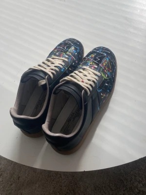 Maison margiela  - Unika svarta sneakers från Maison Margiela med färgstänk i blått, gult, rött och grönt. Skorna har vita snören och beige sula i gummi. Tillverkade i läder med låg profil och klassisk siluett. Perfekta för dig som vill sticka ut med din stil skulle hälst vilja att man köper med instabox, pris kan diskuteras. är kopior.
