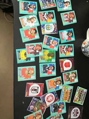 Samling Panini fotbollskort VM 2026 - Säljer en samling Panini fotbollskort från VM 2026. Korten är i färg och visar olika spelare, lag och nationers logotyper. Materialet är glansigt papper och varje kort har en unik design med spelarinformation. Perfekt för dig som gillar att samla på sportkort eller vill byta med vänner.