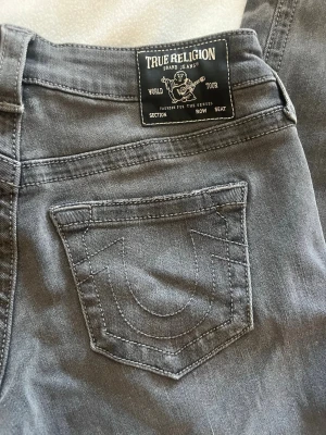Grå jeans från True Religion - Säljer ett par grå jeans från True Religion med klassiska bakfickor och tydliga sömmar. Jeansen har rak passform och är tillverkade i ett mjukt denimtyg. Perfekta för dig som gillar streetwear och vill ha en snygg detalj i garderoben. Modellen heter: SARAH high rise straight 🚫obs! Byxorna är inte högmidjade, utan mer mid/low waist 🚫 storleken är W27, Innerbenslängd 81cm