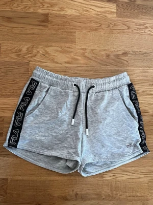 Grå shorts med snörning från FILA - Snygga grå shorts från FILA med svarta snören i midjan och logga på sidan. Shortsens sidor har svarta band med FILA-tryck och de har två fickor fram. Perfekta för en sportig och avslappnad stil.