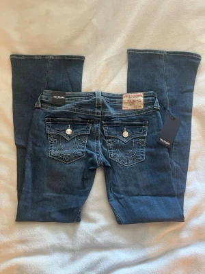 Blå bootcut jeans True Religion - Säljer ett par blå bootcut jeans från True Religion med dekorativa fickor bak och kontrastsömmar. Jeansen har klassisk femficksmodell, snygga detaljer på ficklocken och etikett bak i midjan. Perfekta för dig som gillar en avslappnad men trendig stil. modellen heter JOEY low rise flare 🌟 Storleken är W27, innerbenslängden är 82,5cm 🔥 de är aldrig använda och har kvar prislapp 💫💫