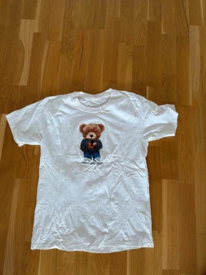 Vit t-shirt med nalletryck - Vit t-shirt i bomull med ett stort tryck av en söt nallebjörn på bröstet. Nallen har på sig en mörk jacka och blå jeans. T-shirten har klassisk passform och rund hals. Perfekt för dig som gillar gulliga och lekfulla motiv. Ser ut som Ralph Lauren teddy. Medium. Oanvänd 