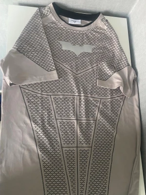 Young LA tränings t shirt i Batman - Snygg Batman cosplaydräkt i stretchigt grått tyg med svart tryckt mönster och Batmansymbol på bröstet. Perfekt för maskerad eller cosplay. Kortärmad modell med detaljer som liknar Batmans rustning. Materialet känns mjukt och följsamt.